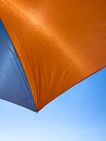 Sitting under the shadow of a blue and orange colored beach umbrella. The shot is taken from below looking up. Fabric of striped umbrella sun protection. Blue sky background with copy space