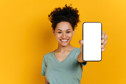 Happy cute african american girl with curly hair, wearing a t-shirt, shows a smartphone in her hand with a blank white screen, stands on an isolated orange background, looks at camera, smiling