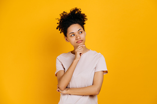 Pensive african american brunette curly haired girl in casual clothes, minded, smart, creative female, look at empty space, thinking, make choice, planning, isolated orange background. Copy-space