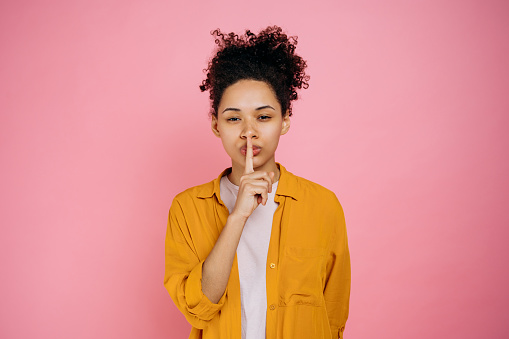 Portrait of a pretty, curly haired african american girl, showing shh sign with finger near lips, silence gesture, secret concept, standing on  isolated pink background, looking at the camera