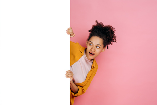 Amazed african american girl with curly hair, peeking out from blank white board with mockup template and copy space for presentation or advertising, stand on pink background, smiles. Mock-up concept