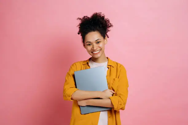 Beautiful positive african american curly haired young woman, wearing casual stylish clothes, holding laptop, standing over isolated pink background, looking at camera, smiling friendly