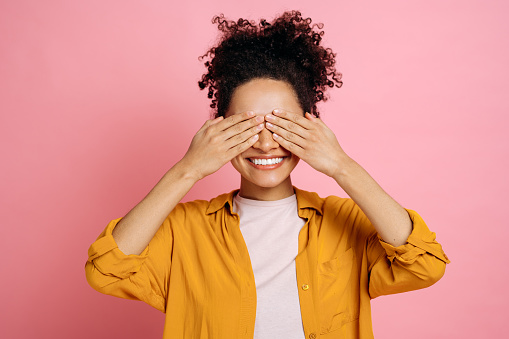 Joyful african american curly haired girl in casual clothes, closes her eyes with her hands, waiting for a surprise, stands on an isolated pink background, smiles happily