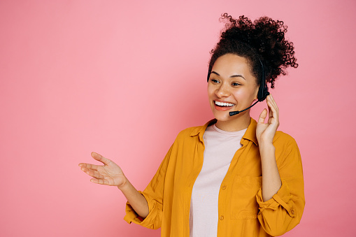 Positive african american curly haired young woman with headset, customer support service, call center operator, expert, advises clients, standing on isolated pink background, smiling friendly