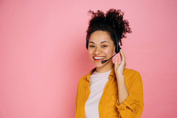 Happy african american curly haired young woman with headset, help desk worker, call center operator, consultant, talking with client, stand on isolated pink background, looks at camera,smile friendly stock photo