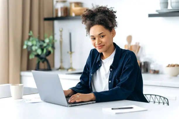 Photo of Portrait of lovely confident african american female student, freelancer, company worker, working or studying remotely, sitting in the kitchen at the table with a laptop, looking at the camera, smiles