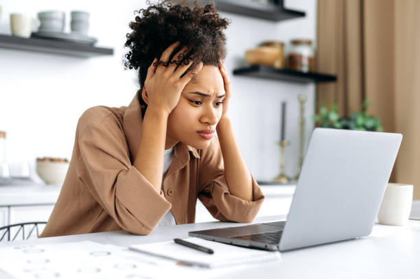 desperate worried sad african american young woman, using laptop for work or study from home, sitting in kitchen, frustrated looking at screen, failed project, got financial loss, job downgrade - downgrade imagens e fotografias de stock