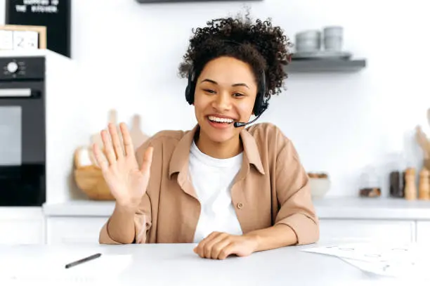 Photo of Positive confident curly-haired young african american woman with headset, call center worker, consultant, talks via video call, female mentor recording webinar, lecture, gesturing with hands, smiling