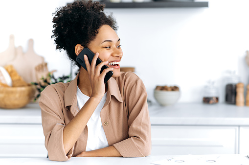 Positive confident african american curly-haired young woman sits at home in the kitchen, talking by smart phone with friend, family or boyfriend, looks away, smiling. Cellphone conversation concept.