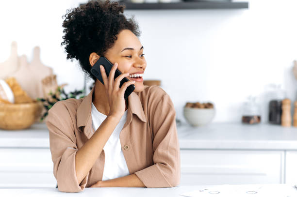 Positive confident african american curly-haired young woman sits at home in the kitchen, talking by smart phone with friend, family or boyfriend, looks away, smiling. Cellphone conversation concept. stock photo