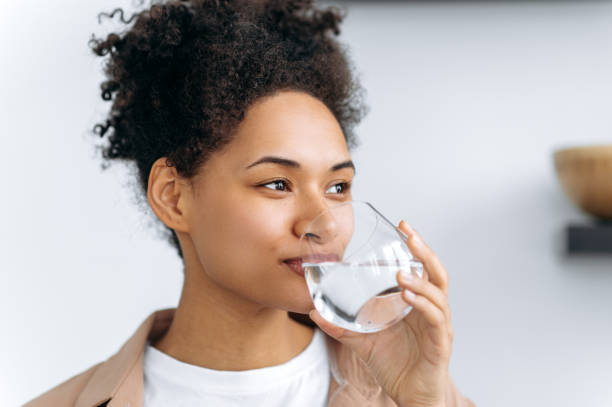 Healthy lifestyle concept. Pretty african american young woman drinks a glass of water at home. Lovely happy healthy mixed race girl follow healthy lifestyle, drink the daily amount of water stock photo