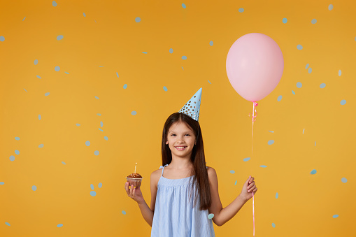 happy little girl with party cone holding cupcake and balloon on her birthday on yellow background with blue confetti