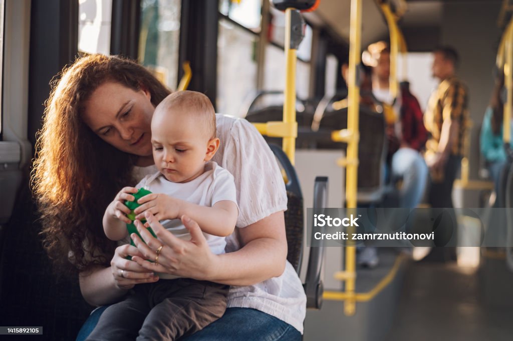 Mother and her child riding in a bus during a day. Young plus size mother in casual wear holding little toddler boy in her lap while riding a bus together in sunny day. Mom and her child enjoying a bus journey together. Bus Stock Photo