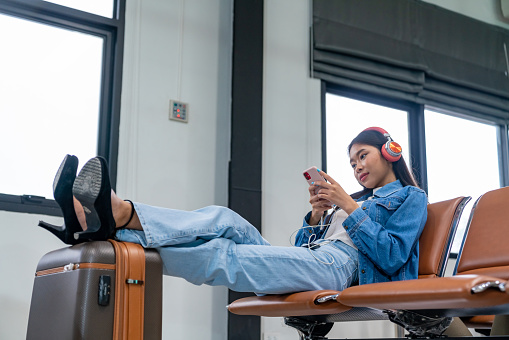 Young Asian woman using mobile phone with internet for online shopping or booking hotel while waiting for boarding time in airport terminal waiting hall. Airplane transportation and holiday vacation concept