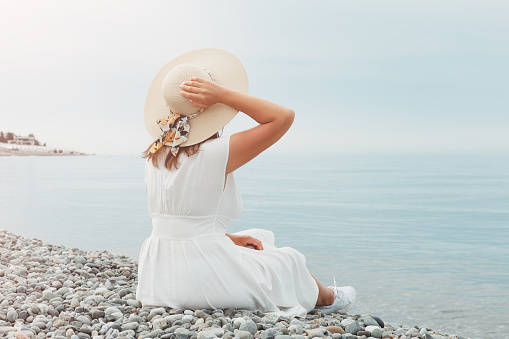 A young girl in a white dress is sitting on the seashore in the rays of the sun, looking into the distance. She holds the straw hat with her hand.