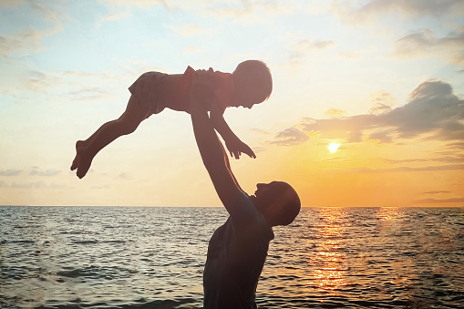The silhouette of a father and child against the background of a sea sunset. A man throws a cheerful, laughing child.