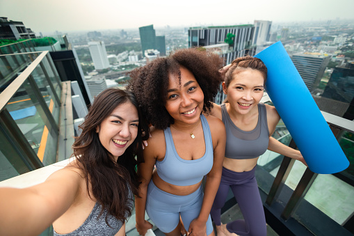 Group of happy fit friends exercising outdoor in city.Smiling young Asian woman looking at African woman play mobile on rooftop city bangkok.