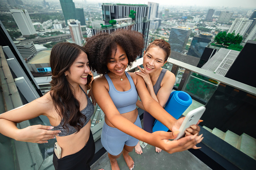 Group of happy fit friends exercising outdoor in city.Smiling young Asian woman looking at African woman play mobile on rooftop city bangkok.