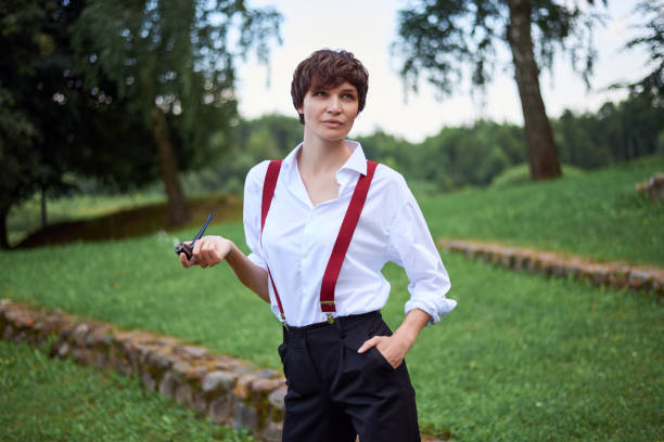 Image of a stylish beautiful woman in a white shirt, black trousers and red suspenders who smokes a pipe. The concept of style and fashion. stock photo