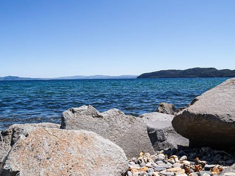 Beautiful blues of sky and lake water beyond rocky water's edge in bay on Lake Taupo, New Zealand.