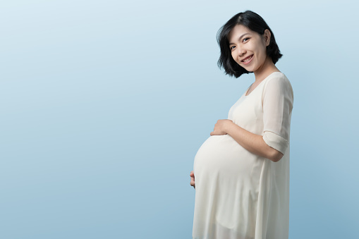 Portrait of Young beautiful Korean or Chinese pregnant woman standing on Blue background, Positive Asian expectant mother over isolated studio wall