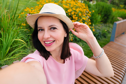 Self portrait brunette woman takes selfie holds camera hand sideways smiles gladfully summer outfit in pastel colors and straw hat outdoors. Caucasian girl posing sitting on bench in park