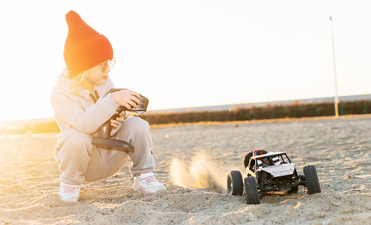 Baby girl playing high speed racing rc car off road buggy vehicle toy radio controller remotely driving. Happy female kid rally safari drive surrounded by sand on black SUV automobile hold joystick