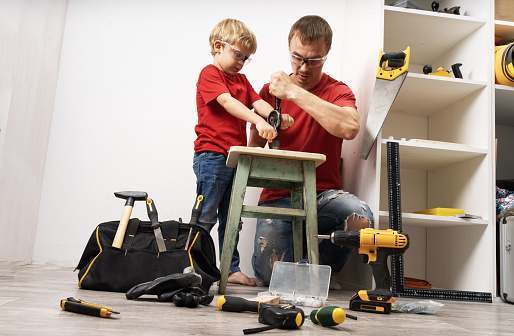Father teaches son to work with a hand drill and other tools. Furniture repair. Home portrait.