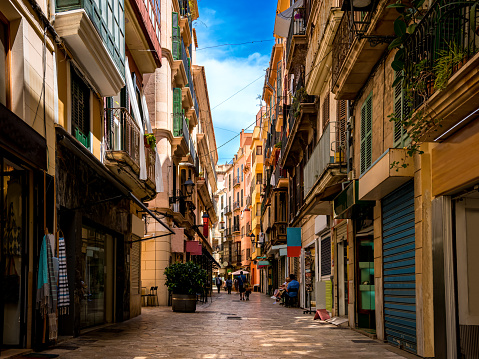 Few tourists walk through a narrow mediterranean alley with small shops and restaurants in the shadow of residential buildings in the groomed centre of Palma de Mallorca on an idyllic day in summer.