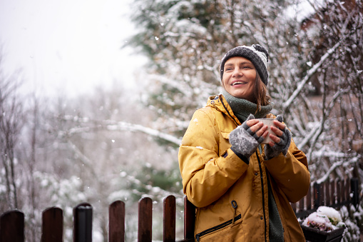 Young happy adult caucasian woman smiling in hat and yellow jacket enjoying tea in a mug with snowfall on the terrace