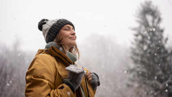 Young adult woman in a hat and yellow jacket breathing fresh air in the winter forest