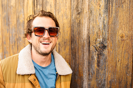 Portrait of a stylish young man wearing sunglasses and a coat laughing while leaning against a wood fence outdoors