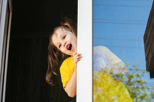 a little cheerful blonde girl in yellow clothes stands on the window of her house and looks out into the street with curiosity