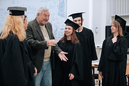 In the math class, an elderly gray-haired professor congratulates students in robes and caps on the completion of their studies and hugs them.