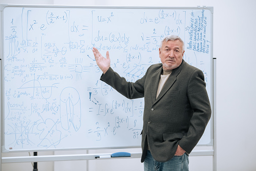 An elderly professor of mathematics stands in his office at a white board with notes and explains the material to students.