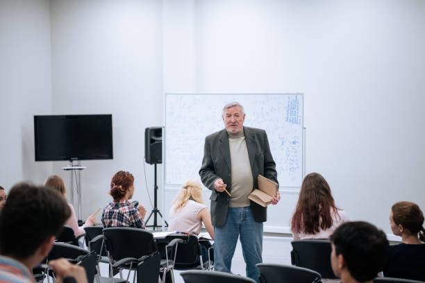 A male teacher in a jacket is giving a lecture on mathematics at the university, he is standing at the white board, students are sitting in a large audience on chairs and listening attentively stock photo