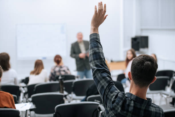 有名な教授が特別なレッスンを行うために大学に到着しました、学生は彼に質問をするために彼の手を引き上げます - 講義室 ストックフォトと画像