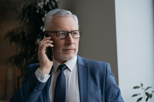 Portrait of pensive senior businessman wearing suit and stylish eyeglasses talking on mobile phone looking through window standing in office