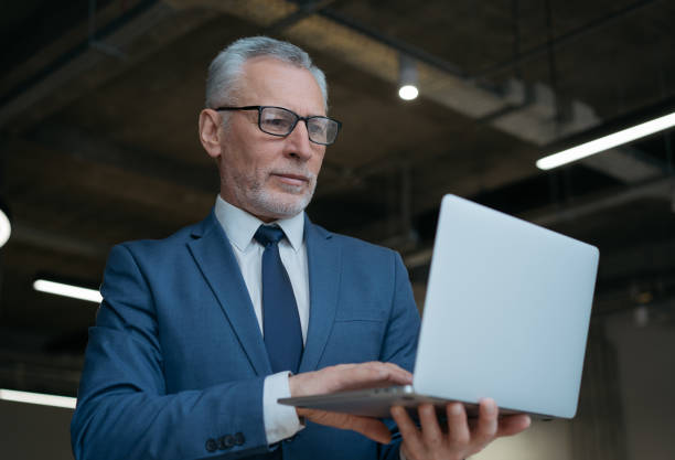 istock Pensive grey haired businessman using laptop computer, typing, searching online, reading news at workplace. Handsome senior man wearing suit and glasses working in modern office 1415963563