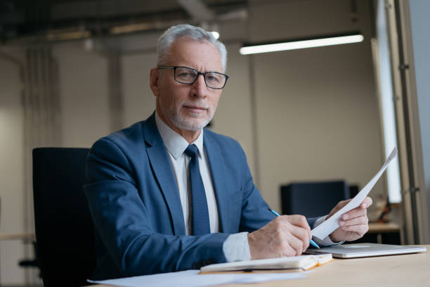 Businessman holding paper documents working in modern office. Portrait of pensive senior manager wearing formal clothing looking at camera sitting at workplace. Successful business stock photo