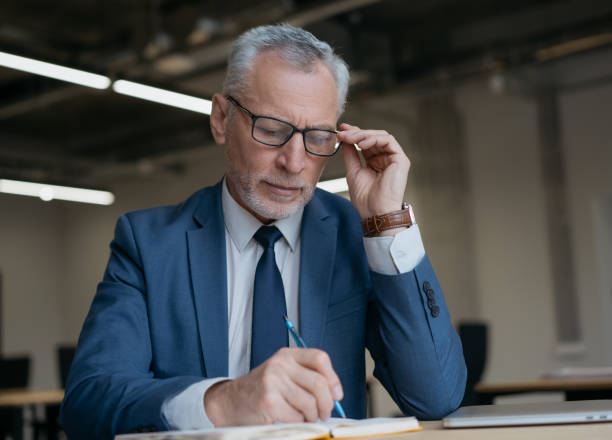 istock Handsome businessman taking notes, planning project, working with financial documents in office. Successful business. Pensive senior manager in eyeglasses writing on paper at workplace 1415964210