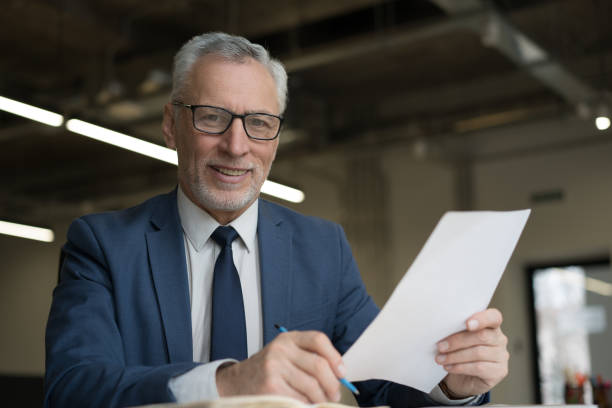 istock Handsome senior businessman holding documents sitting at workplace. Smiling successful manager looking at camera, working in office 1415964287