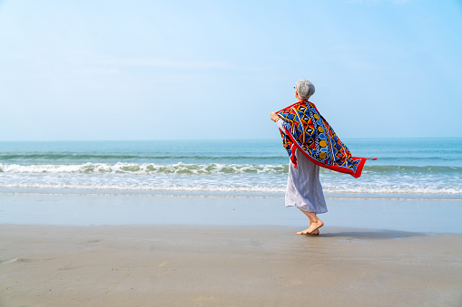 Happy Asian senior woman in blue dress walking on tropical island beach at summer sunset. Elderly retired woman relax and enjoy outdoor lifestyle activity in summer beach holiday travel vacation.