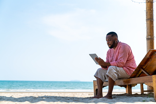 Modern African businessman working on digital tablet for business stocks trading on tropical beach in summer sunny day. Freelancer guy enjoy outdoor lifestyle work and travel on summer travel vacation