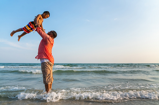 Happy African family on beach holiday vacation. Father playing and carrying little son at tropical beach at summer sunset. Dad with boy kid enjoy and fun outdoor lifestyle activity together at the sea