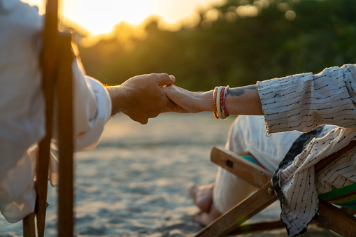 Happy Asian family senior couple enjoy outdoor lifestyle on summer beach holiday vacation at the sea. Elderly retired man and woman relaxing on beach chair and holding hands together at summer sunset