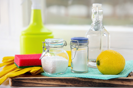 Natural organic eco friendly home cleaning tools ingredients, white vinegar, lemon, baking soda, citric acid on wood tray on window sill, window on background.