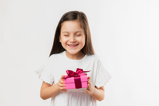 Happy little child girl in a warm mustard knitted sweater holding a red box with a gift on a cyan background. The concept of celebration, giving and receiving a gift.