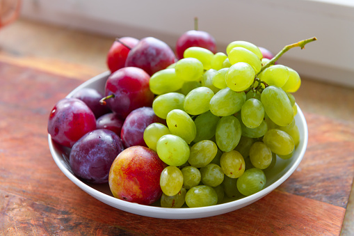 green grapes and plums in a plate on a wooden board, windowsill, concept of fresh fruits and healthy food