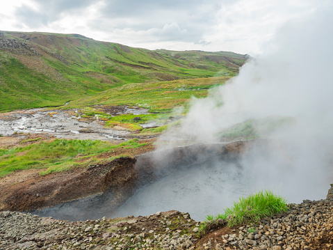 Reykjadalur valley with hot springs river and pool with lush green grass meadow and hills with geothermal steam. South Iceland near Hveragerdi city. Summer sunny morning, blue sky